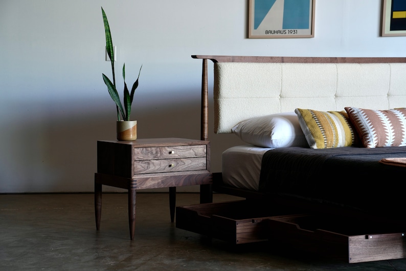 May include: A wooden bed frame with a white tufted headboard and two drawers. The bed has a dark gray blanket and two pillows with geometric patterns. A wooden nightstand with two drawers sits next to the bed, with a potted plant on top.