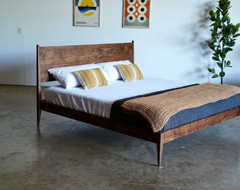 May include: A wooden bed frame with a dark walnut finish. The bed has a headboard with a simple, modern design. The bed is made of solid wood and has a natural wood grain. The bed is dressed with white bedding, a tan throw blanket, and two yellow and white patterned pillows.