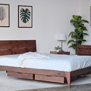 May include: A wooden bed frame with a white mattress and a white blanket. The bed frame has two drawers underneath. The bed is in a room with a white wall and a plant in the corner.