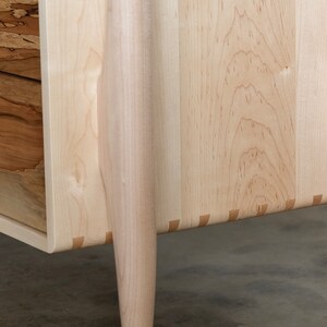 Op de afbeelding: Close-up van de onderkant van een houten dressoir met vier poten. Het dressoir is gemaakt van licht hout met een natuurlijke afwerking. De poten zijn taps toelopend en hebben een licht gebogen vorm. Het dressoir heeft een eenvoudig, elegant ontwerp.