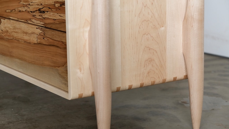 Op de afbeelding: Close-up van de onderkant van een houten dressoir met vier poten. Het dressoir is gemaakt van licht hout met een natuurlijke afwerking. De poten zijn taps toelopend en hebben een licht gebogen vorm. Het dressoir heeft een eenvoudig, elegant ontwerp.