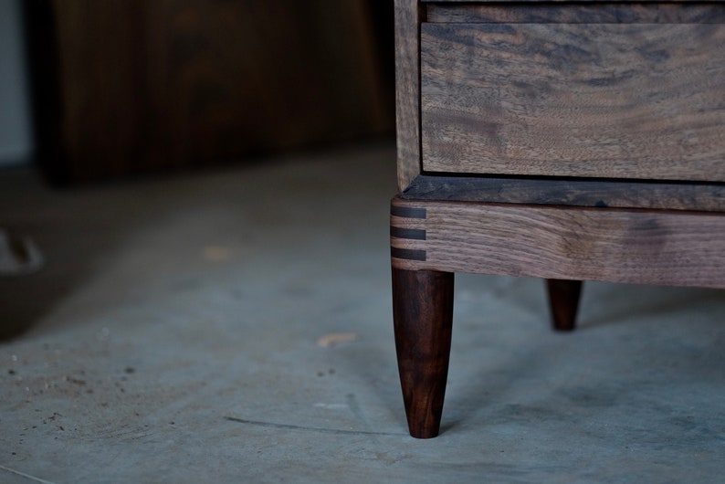 May include: A wooden dresser with a dark brown finish and a drawer. The dresser has four legs with a unique design and a dark brown finish.