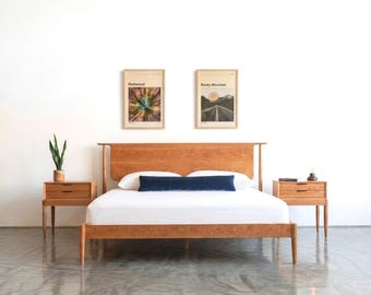 May include: A wooden bed frame with a tall headboard, two matching wooden nightstands, and a white mattress. A long dark blue bolster pillow rests on the bed. Two framed prints hang on the wall above the bed, and a small green plant sits on one nightstand.