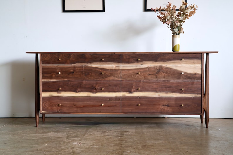 Op de afbeelding: Een dressoir met zes laden van walnoot hout met messing beslag. Het dressoir heeft een mid-century modern design met taps toelopende poten.