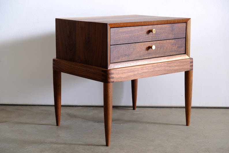 May include: A two-drawer nightstand made of dark wood with brass knobs. The nightstand has four tapered legs and a slightly raised base.