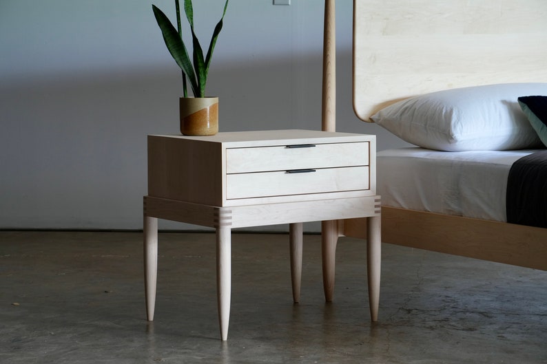 May include: A light wood nightstand with two drawers and four tapered legs. The nightstand is next to a bed with a white headboard and white bedding.
