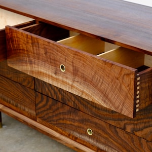 May include: A six-drawer dresser made of dark wood with a wood grain pattern. The top drawer is open, revealing a light wood interior. The dresser has brass drawer pulls and sits on four short legs.