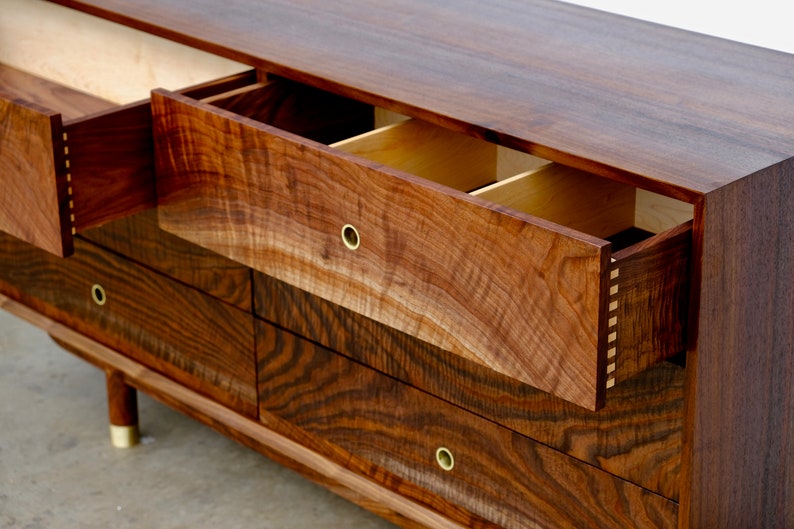 May include: A six-drawer dresser made of dark wood with a wood grain pattern. The top drawer is open, revealing a light wood interior. The dresser has brass drawer pulls and sits on four short legs.