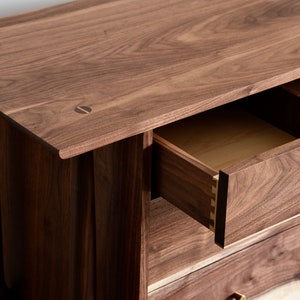 Op de afbeelding: Een houten dressoir met drie lades. De bovenste lade is open, waardoor een licht houten interieur zichtbaar is. Het dressoir is gemaakt van donker hout met een natuurlijke afwerking. De lades hebben messing handgrepen.