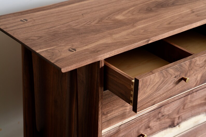 Op de afbeelding: Een houten dressoir met drie lades. De bovenste lade is open, waardoor een licht houten interieur zichtbaar is. Het dressoir is gemaakt van donker hout met een natuurlijke afwerking. De lades hebben messing handgrepen.