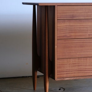 Op de afbeelding: Een houten dressoir met vier lades en messing knoppen. Het dressoir heeft een mid-century modern design met taps toelopende poten.