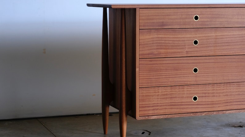Op de afbeelding: Een houten dressoir met vier lades en messing knoppen. Het dressoir heeft een mid-century modern design met taps toelopende poten.