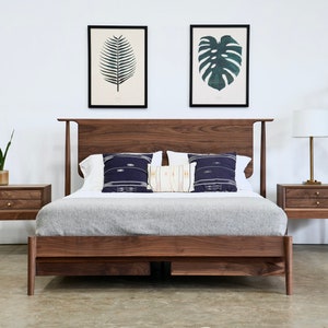 May include: A wooden bed frame with a headboard and footboard. The bed is made of dark wood and has a gray blanket on top. There are two nightstands on either side of the bed, each with two drawers. The nightstands are also made of dark wood. There are two framed prints on the wall behind the bed, one with a fern and the other with a monstera leaf.