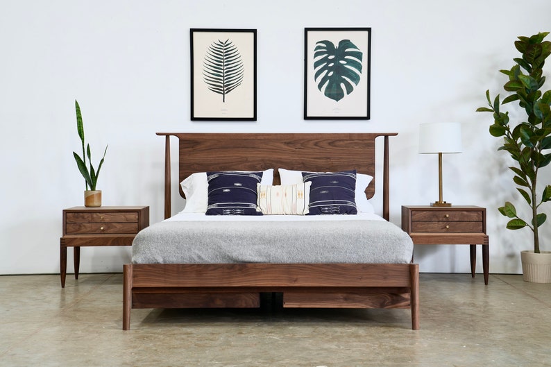 May include: A wooden bed frame with a headboard and footboard. The bed is made of dark wood and has a gray blanket on top. There are two nightstands on either side of the bed, each with two drawers. The nightstands are also made of dark wood. There are two framed prints on the wall behind the bed, one with a fern and the other with a monstera leaf.