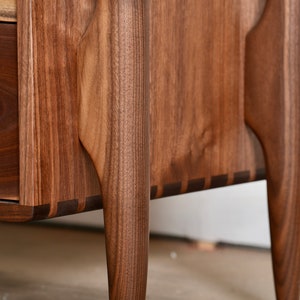 Op de afbeelding: Close-up van een houten dressoir met een lade en taps toelopende poten. Het dressoir is gemaakt van donker hout met een natuurlijke houtnerf. De poten zijn van een donkerdere houtsoort en hebben een uniek, gebogen ontwerp.