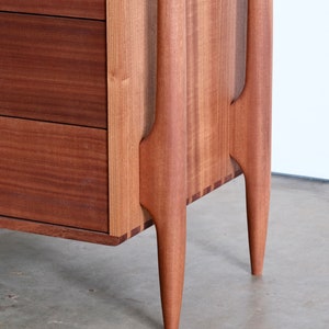 Op de afbeelding: Een houten dressoir met twee laden en unieke, taps toelopende poten. Het dressoir is gemaakt van een rijk, donker hout met een gladde afwerking.