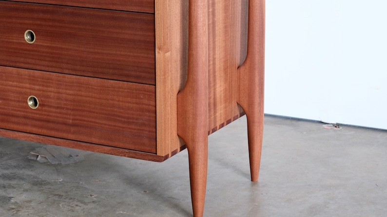 Op de afbeelding: Een houten dressoir met twee laden en unieke, taps toelopende poten. Het dressoir is gemaakt van een rijk, donker hout met een gladde afwerking.