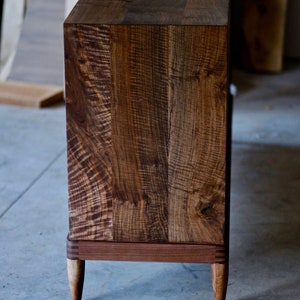 May include: A dark brown wooden cabinet with a natural wood finish. The cabinet has a simple design with a rectangular top and four legs. The legs are tapered and have a slightly curved shape. The cabinet is made of solid wood and has a natural wood grain pattern.