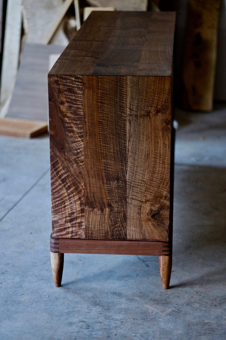 May include: A dark brown wooden cabinet with a natural wood finish. The cabinet has a simple design with a rectangular top and four legs. The legs are tapered and have a slightly curved shape. The cabinet is made of solid wood and has a natural wood grain pattern.