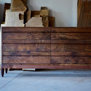 May include: A six-drawer dresser made of dark wood with a rich grain pattern. The dresser has a simple design with straight lines and a slightly curved front. The drawers have a subtle lip around the edges and are set into the frame of the dresser. The dresser is sitting on a concrete floor.