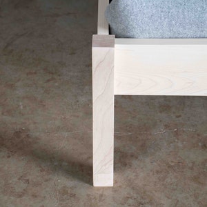 K&ouml;nnte beinhalten: Nahaufnahme eines wei&szlig;en Holzbettgestells mit einem grauen Stoffkopfst&uuml;ck. Das Bettgestell ist aus massivem Holz gefertigt und hat ein schlichtes, modernes Design.