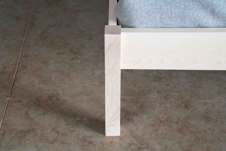 K&ouml;nnte beinhalten: Nahaufnahme eines wei&szlig;en Holzbettgestells mit einem grauen Stoffkopfst&uuml;ck. Das Bettgestell ist aus massivem Holz gefertigt und hat ein schlichtes, modernes Design.