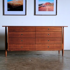 Op de afbeelding: Een dressoir met zes lades van donker hout met messing beslag. Het dressoir heeft een eenvoudig, modern ontwerp uit het midden van de eeuw met taps toelopende poten.