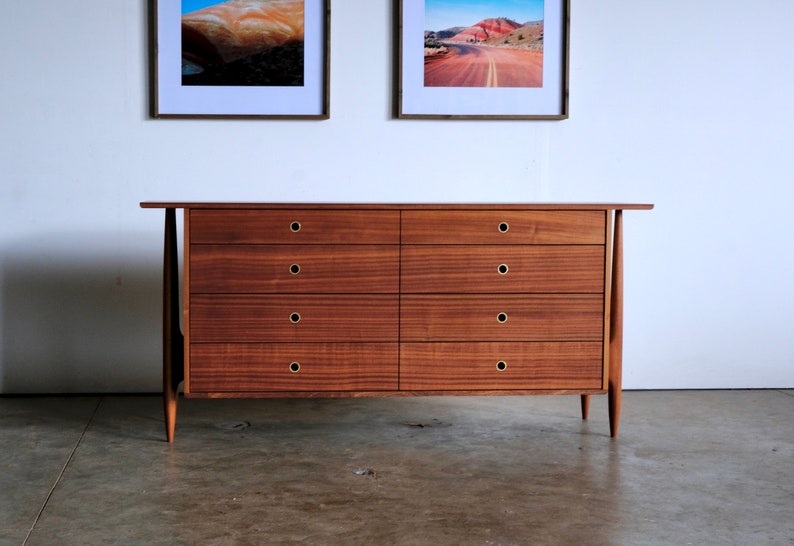 Op de afbeelding: Een dressoir met zes lades van donker hout met messing beslag. Het dressoir heeft een eenvoudig, modern ontwerp uit het midden van de eeuw met taps toelopende poten.