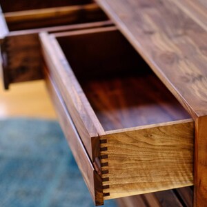 May include: Close-up of a wooden dresser drawer with a dovetail joint. The drawer is open and the wood grain is visible. The dresser is made of walnut wood.