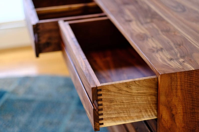 May include: Close-up of a wooden dresser drawer with a dovetail joint. The drawer is open and the wood grain is visible. The dresser is made of walnut wood.