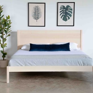 May include: A light wood platform bed frame with a white bedspread and a dark blue lumbar pillow. The bed is in a room with white walls and a potted plant.