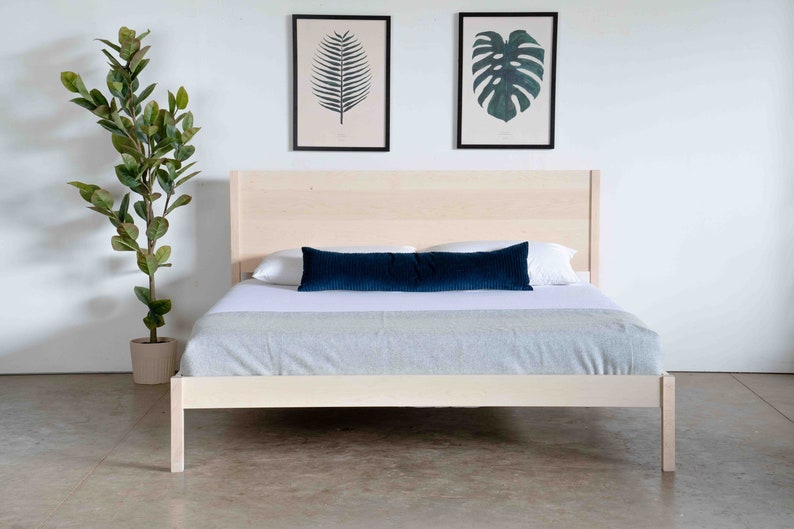 K&ouml;nnte beinhalten: Ein helles Holz-Plattformbettgestell mit einem wei&szlig;en Bettbezug und einem dunkelblauen Lendenkissen. Das Bett steht in einem Raum mit wei&szlig;en W&auml;nden und einer Topfpflanze.
