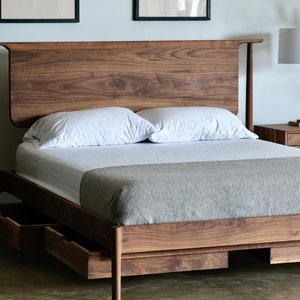 May include: A wooden bed frame with a headboard and footboard. The bed frame has a gray blanket and two white pillows on top. The bed frame has two drawers underneath. The bed frame is in a room with a wooden nightstand and a lamp.