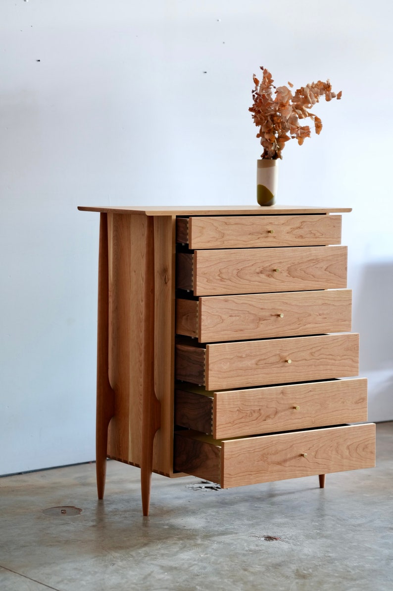Op de afbeelding: Een dressoir met zes lades van licht hout met messing ladegrepen. De lades zijn open, waardoor de binnenkant van het dressoir zichtbaar is. Het dressoir staat op een betonnen vloer.
