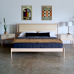 May include: A light wood bed frame with a black headboard and a tan and blue quilt. Two matching light wood nightstands with drawers are on either side of the bed. There are two framed prints on the wall behind the bed.