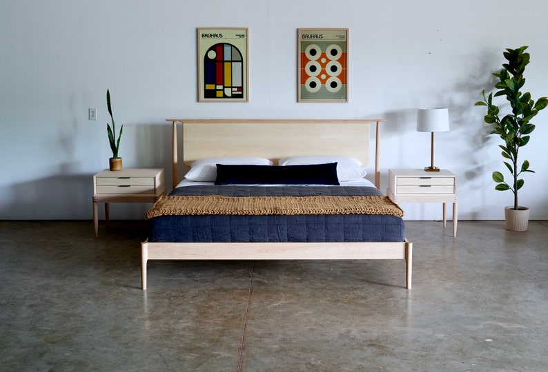 May include: A light wood bed frame with a black headboard and a tan and blue quilt. Two matching light wood nightstands with drawers are on either side of the bed. There are two framed prints on the wall behind the bed.