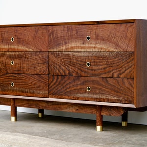 May include: A six-drawer dresser made of dark wood with brass hardware and rounded legs. The dresser has a wood grain pattern and is made of solid wood.