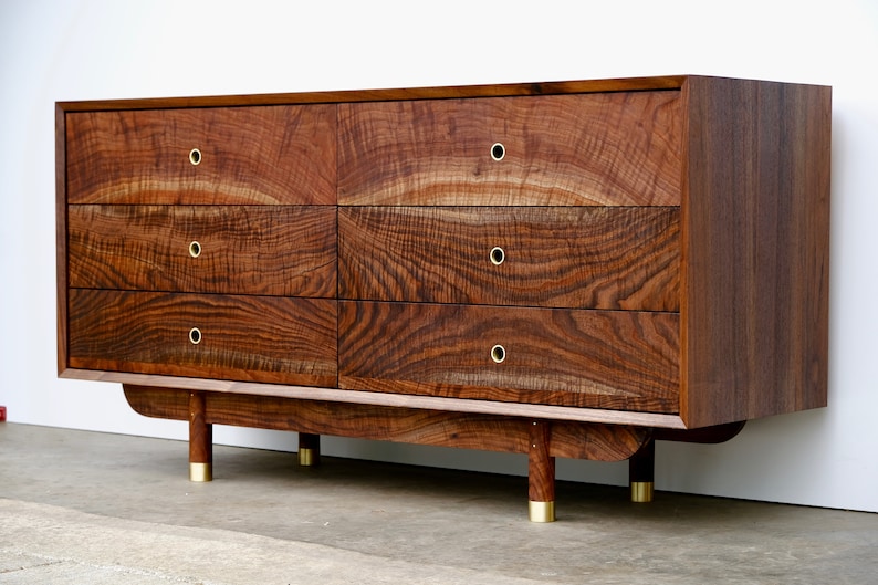 May include: A six-drawer dresser made of dark wood with brass hardware and rounded legs. The dresser has a wood grain pattern and is made of solid wood.