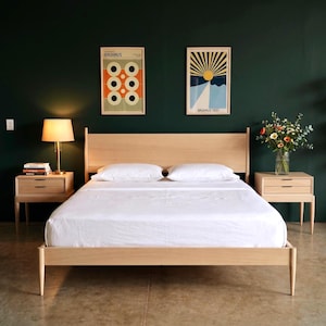 May include: A light wood bed frame with a headboard, two nightstands, and a white bedspread. Two framed Bauhaus art prints hang on the dark green wall. A lamp sits on one nightstand, and a vase of flowers on the other.