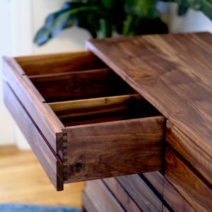 May include: A wooden dresser with multiple drawers, one of which is open, revealing a dark wood interior with dividers. The dresser is made of a rich, dark wood with a smooth finish.