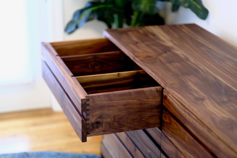 May include: A wooden dresser with multiple drawers, one of which is open, revealing a dark wood interior with dividers. The dresser is made of a rich, dark wood with a smooth finish.