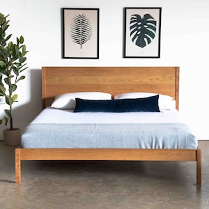 May include: A wooden bed frame with a light wood finish. The bed has a headboard and footboard, and a blue throw blanket is on the bed. There are two framed prints on the wall behind the bed, one with a green leaf and one with a monstera leaf.
