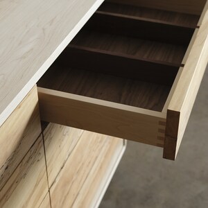 Op de afbeelding: Een houten dressoir met een open lade, die een licht houten interieur met verdelers onthult. Het dressoir is gemaakt van licht en donker hout.