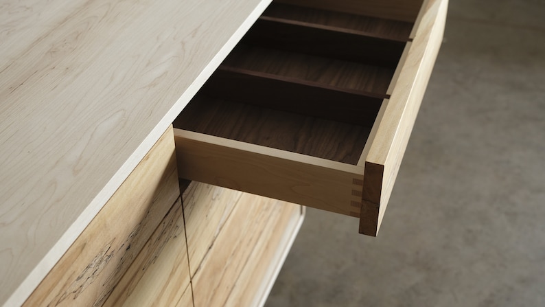 Op de afbeelding: Een houten dressoir met een open lade, die een licht houten interieur met verdelers onthult. Het dressoir is gemaakt van licht en donker hout.