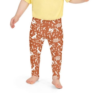 May include: A toddler wearing a yellow t-shirt and brown leggings with a white pattern of animals and plants.