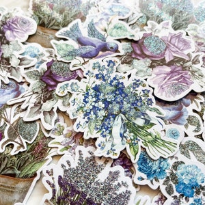 K&ouml;nnte beinhalten: Eine Sammlung von Blumenaufklebern im Vintage-Stil mit glitzernder Oberfl&auml;che. Die Aufkleber zeigen verschiedene Blumen, darunter Rosen, Vergissmeinnicht und Lavendel, in Blaut&ouml;nen, Lila und Gr&uuml;n.