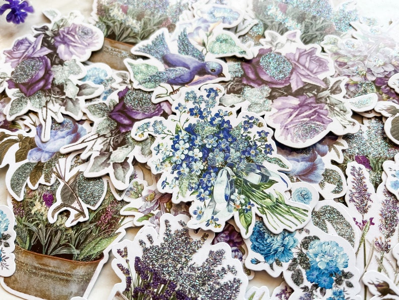 K&ouml;nnte beinhalten: Eine Sammlung von Blumenaufklebern im Vintage-Stil mit glitzernder Oberfl&auml;che. Die Aufkleber zeigen verschiedene Blumen, darunter Rosen, Vergissmeinnicht und Lavendel, in Blaut&ouml;nen, Lila und Gr&uuml;n.