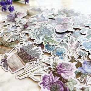 K&ouml;nnte beinhalten: Eine Sammlung von Aufklebern mit Lavendel- und Blumendesigns mit Glitzerakzenten. Die Aufkleber zeigen verschiedene Designs, darunter Blumenstr&auml;u&szlig;e, einzelne Blumen und einen Blumentopf.