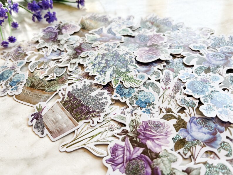 K&ouml;nnte beinhalten: Eine Sammlung von Aufklebern mit Lavendel- und Blumendesigns mit Glitzerakzenten. Die Aufkleber zeigen verschiedene Designs, darunter Blumenstr&auml;u&szlig;e, einzelne Blumen und einen Blumentopf.