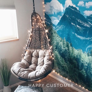 Puede incluir: Una silla colgante de mimbre marr&oacute;n con un coj&iacute;n beige, decorada con luces de cadena. La silla est&aacute; colgada frente a un tapiz que representa una escena de monta&ntilde;a con cielo azul y &aacute;rboles verdes.